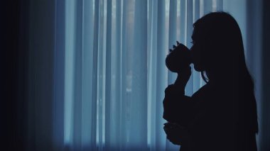 Yumuşak ışıklı bir pencerenin önünde sıcak bir içkinin keyfini çıkaran bir kadının silüeti, sakinlik ve rahatlama iletiyor..