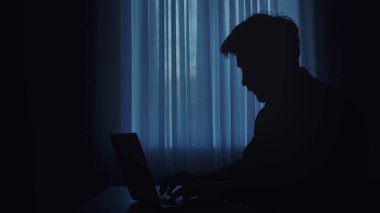 Silüetli bir kişi karanlık bir odada dizüstü bilgisayarda çalışır. Pencereden gelen yumuşak mavi ışıkla aydınlatılır..