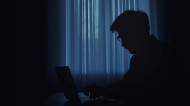 Bir insan, bir dizüstü bilgisayarın parıltısına karşı siluetlenir, loş bir alanda yazar, gizemli bir atmosfer yaratır..