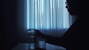 Pencerenin kenarında oturan bir kişinin silueti masanın üzerindeki bir su bardağına uzanıyor, sakin ve düşünceli bir atmosfer yaratıyor..