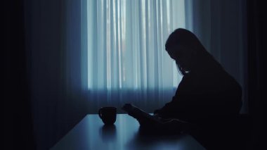 Bir kadın silueti pencerenin kenarında oturur, sakin, loş bir odada kitap okur. Atmosfer sakin ve dalgın..