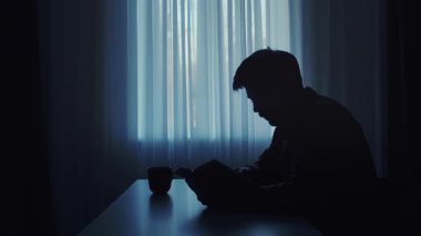 Pencerenin yanında kitap okuyan bir kişinin silueti huzur verici ve iç gözlem sahnesi yaratıyor..