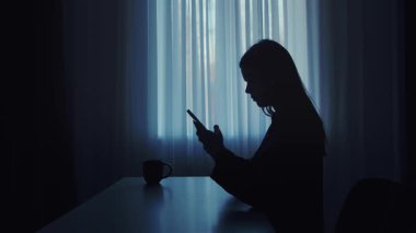 Pencerenin önündeki masada oturan bir kadının silüeti. Yanında bardakla akıllı telefon kullanıyor..