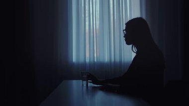 Pencerenin kenarında oturan bir kişinin silüeti, bir bardak su tutarak, sakin ve yansıtıcı bir atmosfer yaratıyor..