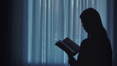 Yumuşak ışıklandırılmış bir arkaplan karşısında kitap okuyan bir kişinin silueti, huzurlu bir anı yakalıyor..