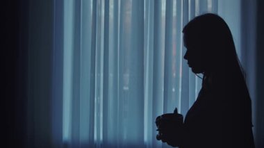 Yumuşak ışıklı bir pencerenin önünde sıcak bir içkinin keyfini çıkaran bir kadının silüeti, sakinlik ve rahatlama iletiyor..
