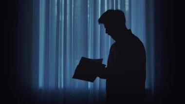 Mavi perdeli bir pencerenin önünde kitap okuyan bir kişinin silueti, sakin ve iç gözlemci bir atmosfer yaratıyor..