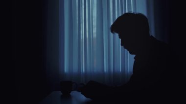 Gizemli bir görüntü perdeli bir pencerenin kenarında oturan bir adamın siluetini gösteriyor, yalnızlık ve tefekküre işaret ediyor. Sahne loş bir ışıkla çevrelenmiş ışığa karşı yumuşak ana hatları vurguluyor.