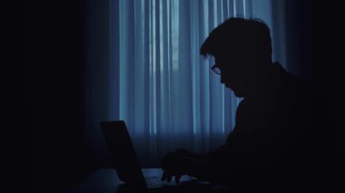 Loş bir odada dizüstü bilgisayarda yazı yazan bir kişinin silüeti gizemli bir atmosfer yaratıyor..