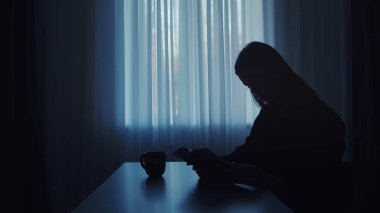 Perdeleri ve kahve fincanıyla pencerenin yanında kitap okuyan bir kadının silueti, huzurlu bir atmosfer yaratıyor..