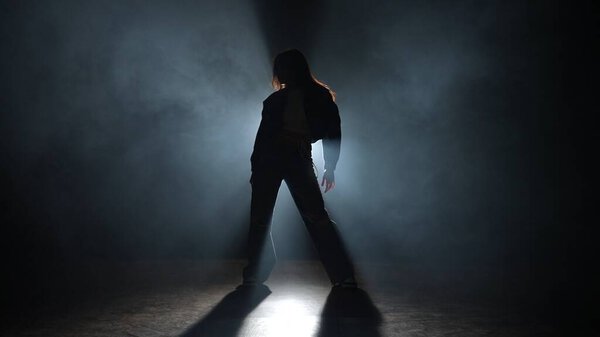 Modern choreography creative concept. Silhouette of a woman dancing jazz-funk in a studio. Black background filled with smoke, white spotlight shining from behind.