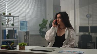 Yaratıcı iş konsepti. Genç iş kadını yönetici ofiste masa başında oturuyor, çalışıyor, telefonlara cevap veriyor, akıllı telefondan konuşuyor, mutlu bir ifade takınıyor. Yavaş çekim.