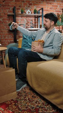 Abur cubur konsepti. Fast food teslimatçısıyla evde vakit geçiren bir adam, elinde tavuk budu, elinde kovayla kanepede oturur ve akıllı telefondan selfie çeker..