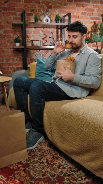 Abur cubur konsepti. Adam evde fast food dağıtımı ile vakit geçiriyor, kanepede oturup televizyon izliyor, kızarmış tavuk bacağını sosa batırıyor ve onu ısırıyor, tadını çıkarıyor..