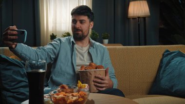 Abur cubur konsepti. Fast food teslimatçısıyla evde vakit geçiren bir adam, elinde tavuk budu, elinde kovayla kanepede oturur ve akıllı telefondan selfie çeker..