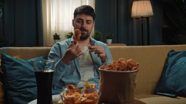 Abur cubur konsepti. Fast food teslimatçısıyla evde vakit geçiren bir adam, elinde tavuk budu, elinde kovayla kanepede oturur ve akıllı telefondan selfie çeker..