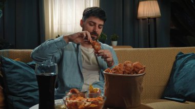 Abur cubur konsepti. Fast food teslimatçısıyla evde vakit geçiren bir adam, elinde tavuk budu, elinde kovayla kanepede oturur ve akıllı telefondan selfie çeker..