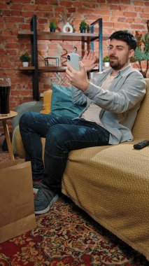 Abur cubur reklamı konsepti. Sakallı adam evde fast food dağıtımı ile vakit geçiriyor, kanepede oturuyor, akıllı telefondan video konuşmaları yapıyor ve kola içiyor..