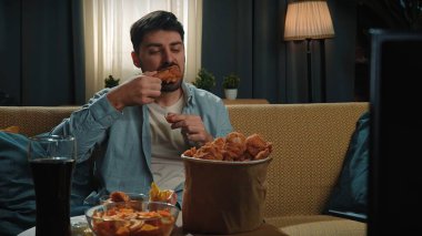 Abur cubur konsepti. Fast food teslimatçısıyla evde vakit geçiren bir adam, elinde tavuk budu, elinde kovayla kanepede oturur ve akıllı telefondan selfie çeker..