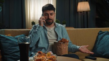 Abur cubur reklamı konsepti. Fast food teslimatçısıyla evde vakit geçiren, kanepede oturup tavuk budu yiyen ve akıllı telefondan konuşan bir adam..