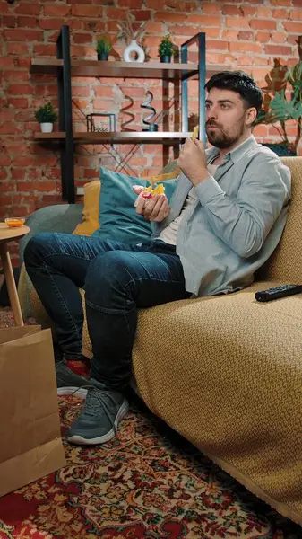 Abur cubur reklamı konsepti. Sakallı adam evde fast food dağıtımı ile vakit geçiriyor, kanepede oturup TV 'de film izliyor, gülümsüyor ve soslu kızarmış patates yiyor..