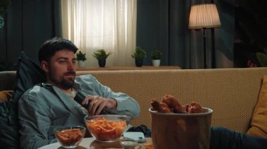 Abur cubur reklamı konsepti. Sakallı adam evde fast food dağıtımıyla vakit geçiriyor, kanepeye uzanıyor, televizyon izliyor, patates cipsi yiyor ve akıllı telefon çeviriyor. Yavaş çekim