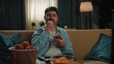 Abur cubur reklamı konsepti. Sakallı adam evde fast food teslimatlarıyla vakit geçiriyor, kanepede oturup televizyon izliyor ve bir kaseden kızarmış patates yiyor, sosa batırıyor. Yavaş çekim