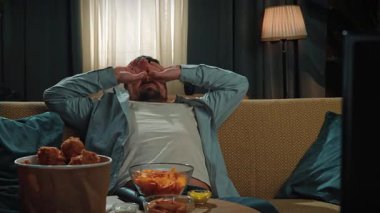 Abur cubur reklamı konsepti. Sakallı adam evde fast food teslimatlarıyla vakit geçiriyor, koltukta uyuyor, uyanıyor ve esneyerek tavuk yemeye başlıyor. Yavaş çekim