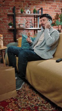 Abur cubur reklamı konsepti. Fast food teslimatçısıyla evde vakit geçiren, kanepeye oturup tavuk budu yiyen ve akıllı telefondan konuşan bir adam. Yavaş çekim, dikey video.