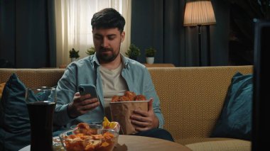 Abur cubur reklamı konsepti. Sakallı adam evde fast food dağıtımı ile vakit geçiriyor, elinde kovayla kanepede oturuyor tavuk budu ve akıllı telefondan mesaj yazıyor..