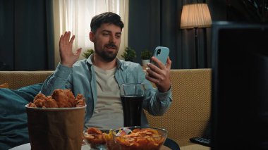 Abur cubur reklamı konsepti. Sakallı adam evde fast food dağıtımı ile vakit geçiriyor, kanepede oturuyor, akıllı telefondan video konuşmaları yapıyor ve kola içiyor..