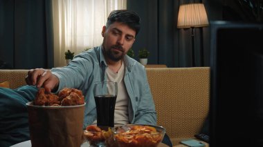 Abur cubur reklamı konsepti. Adam evde fast food dağıtımı ile vakit geçiriyor, kanepede oturup televizyon izliyor, sosa batırılmış tavuk yiyor, kola içiyor, tadını çıkarıyor..