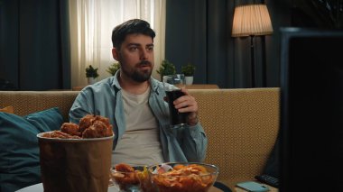 Abur cubur reklamı konsepti. Adam evde fast food dağıtımı ile vakit geçiriyor, kanepede oturup televizyon izliyor, sosa batırılmış tavuk yiyor, kola içiyor, tadını çıkarıyor..