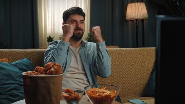 Neşeli bir adam kanepede otururken heyecandan yumruklarını kaldırmış televizyon izler. Önünde bir kase cips, kızarmış tavuk ve atıştırmalıklar var. Sıcak ışıklandırmalı rahat ev ayarları.