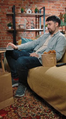 Abur cubur reklamı konsepti. Sakallı adam evde fast food dağıtımı ile vakit geçiriyor, kanepede oturuyor dizüstü bilgisayarında videolar izliyor ve kovadan kızarmış tavuk yiyor..