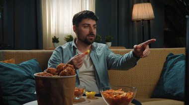 Abur cubur konsepti. Adam evde fast food dağıtımı ile vakit geçiriyor, kanepede oturup televizyon izliyor, kızarmış tavuk bacağını sosa batırıyor ve onu ısırıyor, tadını çıkarıyor..