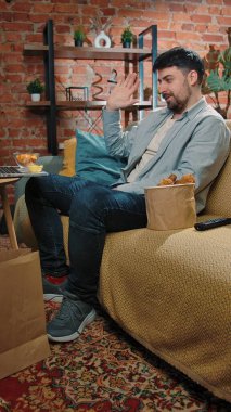 Abur cubur reklamı konsepti. Sakallı adam evde fast food dağıtımı ile vakit geçiriyor, kanepeye oturup bilgisayardan video görüşmesi yapıyor ve soslu kızarmış patates yiyor..