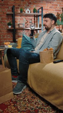 Abur cubur reklamı konsepti. Adam evde fast food dağıtımı ile vakit geçiriyor, kanepede oturup televizyon izliyor, sosa batırılmış tavuk yiyor, kola içiyor, tadını çıkarıyor..