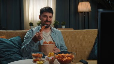 Abur cubur konsepti. Adam evde fast food dağıtımı ile vakit geçiriyor, kanepede oturup televizyon izliyor, kızarmış tavuk bacağını sosa batırıyor ve onu ısırıyor, tadını çıkarıyor..