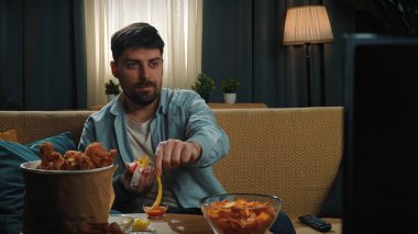 Abur cubur konsepti. Adam evde fast food dağıtımı ile vakit geçiriyor, kanepede oturup televizyon izliyor, kızarmış tavuk bacağını sosa batırıyor ve onu ısırıyor, tadını çıkarıyor..