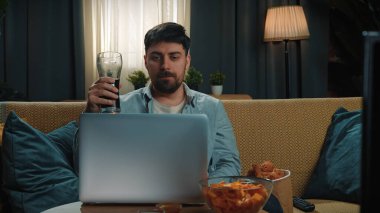 Abur cubur reklamı konsepti. Sakallı adam evde fast food dağıtımı ile vakit geçiriyor, kanepede oturuyor dizüstü bilgisayarında videolar izliyor ve kovadan kızarmış tavuk yiyor..