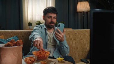 Abur cubur reklamı konsepti. Sakallı adam evde fast food dağıtımı ile vakit geçiriyor, kanepeye oturuyor, televizyon izliyor, patates cipsi yiyor ve akıllı telefonları kaydırıyor..