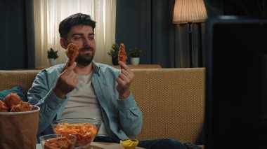 Abur cubur reklamı konsepti. Sakallı adam evde fast food teslimatlarıyla vakit geçiriyor, kanepede oturup televizyon seyrediyor, arkadaşının elini tutuyor, kokluyor ve ısırıyor, tadını çıkarıyor..