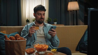 Abur cubur reklamı konsepti. Sakallı adam evde fast food dağıtımı ile vakit geçiriyor, kanepeye oturuyor, pirzola yiyor ve internetten alışveriş yapıyor, akıllı telefondan kart numarası yazıyor..