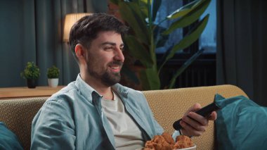 Abur cubur konsepti. Adam evde fast food dağıtımı ile vakit geçiriyor, kanepede oturup televizyon izliyor, kızarmış tavuk bacağını sosa batırıyor ve onu ısırıyor, tadını çıkarıyor..