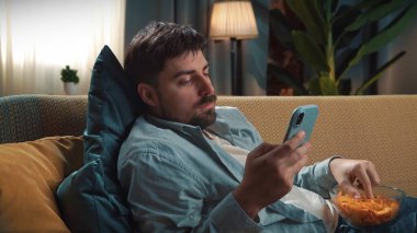 Abur cubur reklamı konsepti. Sakallı adam evde fast food dağıtımı ile vakit geçiriyor, elinde kovayla kanepede oturuyor tavuk budu ve akıllı telefondan mesaj yazıyor..