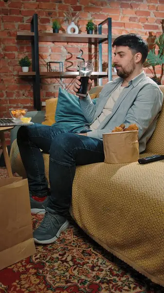 Abur cubur reklamı konsepti. Adam evde fast food dağıtımı ile vakit geçiriyor, kanepede oturup televizyon izliyor, sosa batırılmış tavuk yiyor, kola içiyor, tadını çıkarıyor..