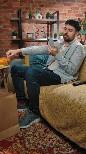 Abur cubur reklamı konsepti. Sakallı adam evde fast food dağıtımı ile zaman geçiriyor, kanepeye uzanıyor, televizyon izliyor, patates cipsi yiyor ve akıllı telefonları kaydırıyor..