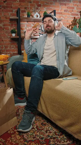 Abur cubur reklamı konsepti. Sakallı adam evde fast food dağıtımı ile vakit geçiriyor, kanepeye oturuyor kulaklıkla müzik dinliyor ve kızarmış tavuk yiyor..