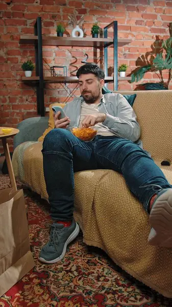 Abur cubur reklamı konsepti. Sakallı adam evde fast food dağıtımı ile zaman geçiriyor, kanepeye uzanıyor, televizyon izliyor, patates cipsi yiyor ve akıllı telefonları kaydırıyor..